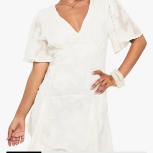 Studentklänning NY MED PRISLAPP - Supersöt vit kortklänning från Boohoo med omlott-design och v-ringning. Klänningen har korta, vida ärmar och ett diskret blommigt mönster i tyget. Materialet är lätt och ger en flowy känsla, perfekt för sommarens alla tillfällen. Snygg detalj med öppen rygg. OBS! Modellen är liten i storlek å sitter som en 38/M. Klänningen är aldrig använd. Nypris 685