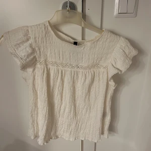 Vit blus med spetsdetalj från H&M - HELT NY, LAPPEN ÄR KVAR! Säljer en söt vit blus från H&M med korta volangärmar och en fin spetsdetalj över bröstet. Blusen är tillverkad i ett krinklat bomullstyg som ger en avslappnad och luftig känsla. Perfekt för vår och sommar! 