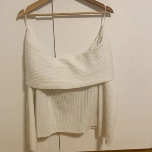 Vit ribbad offshoulder stickad topp - Säljer en vit stickad topp med ribbad struktur och bred offshoulder-krage. Toppen har långa ärmar och en mjuk, cozy känsla som passar perfekt för kyliga dagar. Snygg att styla med jeans eller kjol för en trendig look.