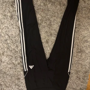 Svarta Adidas träningsbyxor slim fit - Svarta träningsbyxor från Adidas i slim fit med vita ränder längs benen och Adidas-logga på sidan. Byxorna har elastisk midja och är gjorda i polyester, perfekta för sport eller chill. Snygg och sportig look med klassiska detaljer. Aldrig använda