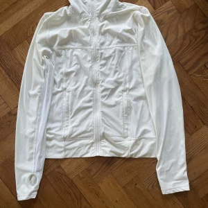 Vit vindjacka från Drop of Mindfulness - Snygg vit vindjacka från Drop of Mindfulness i storlek M. Jackan har dragkedja framtill, två fickor och tumhål vid ärmsluten. Tillverkad i mjuk och stretchig polyamid och elastan, perfekt för träning eller chill. Diskret logga baktill.