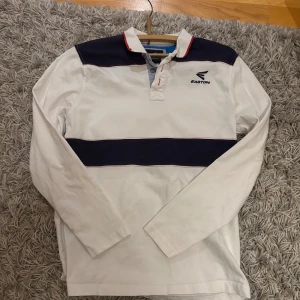 Vit och marinblå pikétröja Easton S - Säljer en vit och marinblå långärmad pikétröja från Easton. Tröjan har breda marinblå ränder över bröst och rygg, klassisk krage med röd detalj och knäppning framtill. Perfekt för dig som gillar sportig och stilren look.