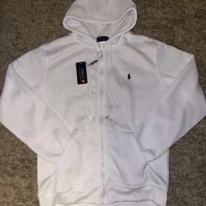 Vit hoodie från Polo Ralph Lauren - Vit hoodie från Polo Ralph Lauren med klassisk svart logobrodyr på bröstet. Tröjan har huva med snörning, dragkedja framtill och två stora fickor. Materialet är mjukt och känns skönt mot huden, perfekt för chill dagar. Snygg och enkel design som alltid funkar.