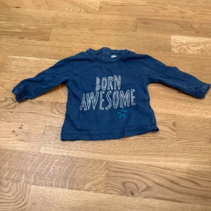 Blå långärmad tröja Babaluno - Supersöt blå långärmad tröja från Babaluno med trycket 'BORN AWESOME' och en liten stjärna framtill. Tröjan har praktiska tryckknappar vid axeln för enkel påklädning. Perfekt för små coola kids!