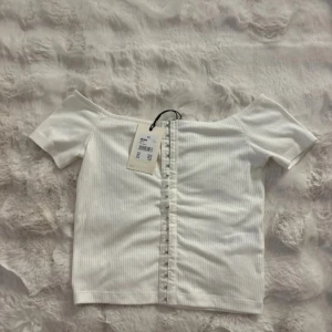 Vit ribbad offshoulder topp - Säljer en fräsch vit ribbad topp från Bershka med offshoulder-design och korta ärmar. Toppen har en snygg rynkad detalj framtill som ger extra stilpoäng. Perfekt för dig som gillar trendiga och enkla plagg med twist.