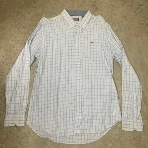 Vit rutig skjorta från Tommy Hilfiger - Vit skjorta från Tommy Hilfiger med ljusblå rutor och klassisk krage. Skjortan har långa ärmar, knappar framtill och ett litet broderat logomärke på bröstet. Tillverkad i mjuk bomull som känns skön mot huden. Perfekt för dig som gillar stilrena och fräscha plagg.