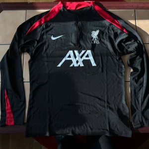  Liverpool träningströja  - Liverpool Half-zip träningströja i svart färg. Ärmarna har hål där tummarna får plats