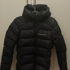 Svart dunjacka Peak Performance - Svart dunjacka från Peak Performance med huva och dragkedja framtill. Jackan har ett puffigt, quiltat utseende och logga på bröstet. Perfekt för kalla dagar med sin tjocka och isolerande design. Snygg och funktionell för dig som gillar stil och värme.