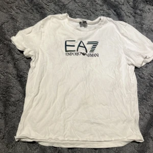 Vit EA7 Emporio Armani t-shirt - Vit t-shirt från EA7 Emporio Armani med stort, färgglatt logotryck på bröstet. Klassisk rund hals och korta ärmar. Mjuk och skön i bomull, perfekt för dig som gillar stilrena märkesplagg med lite extra attityd.