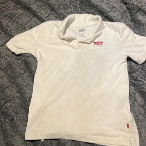 Vit pikétröja från Levi's - Vit pikétröja från Levi's med klassisk krage och korta ärmar. Tröjan har en röd broderad Levi's-logga på bröstet och knappar vid halsen. Materialet är bomull och ger en skön känsla. Perfekt för dig som gillar enkel och stilren design.