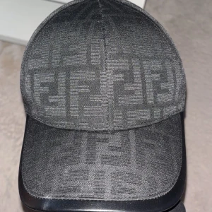 Svart keps från Fendi med FF-mönster - Snygg svart keps från Fendi med det klassiska FF-logomönstret över hela kepsen. Stilren och modern design som passar perfekt till streetwear. Materialet känns mjukt och exklusivt, och kepsen har böjd skärm för en cool look.