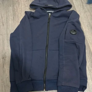 Mörkblå hoodie från Armani Exchange - Snygg mörkblå hoodie från Armani Exchange med dragkedja framtill och huva. Tröjan har lång ärm, ribbade muddar och en cool detalj på ärmen. Perfekt för dig som gillar stilren streetwear.
