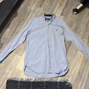 Blå skjorta från Polo Ralph Lauren - Klassisk ljusblå skjorta från Polo Ralph Lauren med ikonisk broderad logga på bröstet. Skjortan har lång ärm, knappar framtill och en snygg krage. Tillverkad i mjuk bomull som känns skön mot huden. Perfekt för dig som gillar stilrena och tidlösa plagg.