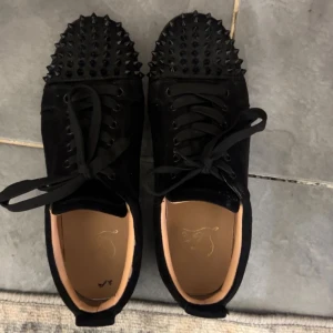 Christian Louboutin svarta sneakers med nitar - Svarta sneakers från Christian Louboutin med coola svarta nitar över tån och klassisk röd sula med logga. Skorna har snörning och är tillverkade i mocka med rund tå och platt sula. Perfekt för dig som vill sticka ut med en edgy och lyxig stil.