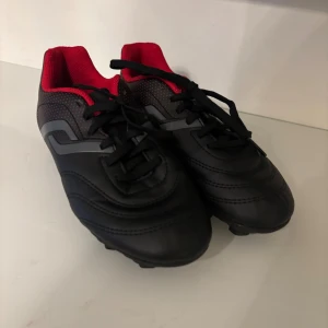 Barn Svarta fotbollsskor med röd insida - Snygga svarta fotbollsskor med röd insida och svarta snören. Skorna har en rund tå och platt sula med dobbar för bra grepp på planen. Sidan har diskreta grå detaljer och små röda prickar. Tillverkade i syntetmaterial som tål tuffa matcher.