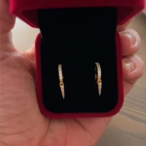 Glittriga guldörhängen med stenar - Snygga örhängen i guldton med glittriga stenar längs hela bågen. De har en smal, spetsig form som ger en modern och cool vibe. Perfekta för dig som gillar att sticka ut med detaljer som verkligen syns. Guldpläterade i 14k. Röda boxen ingår INTE!