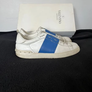 Valentino Garavani sneakers vit/blå - Snygga Valentino Garavani sneakers i vitt skinn med blåa detaljer på sidorna och tungan. Klassisk rund tå, snörning och platt sula med nitar baktill. Perfekt för dig som gillar exklusiva och stilrena sneakers med en cool touch. Dom är rätt så använda. Hör av dig om du har några frågor