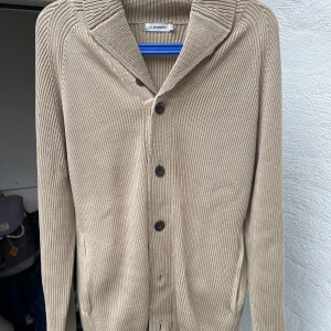 Beige stickad kofta från J.Lindeberg - Snygg beige stickad kofta från J.Lindeberg med ribbad struktur och klassisk krage. Koftan har långa ärmar, knappar framtill och två fickor. Perfekt för lager-på-lager och chill stil. Passar dig som gillar enkel och clean design.