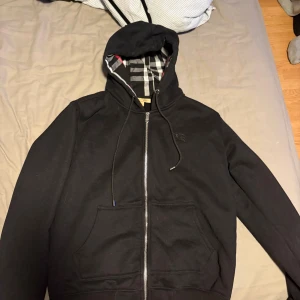 Svart Burberry hoodie med dragkedja - Svart hoodie från Burberry med klassiskt rutigt foder i huvan och broderad logga på bröstet. Tröjan har dragkedja framtill, snörning vid huvan och två stora fickor. Materialet är mjuk bomull med lite elastan för extra komfort. Perfekt för dig som gillar stilren streetwear.Helt ny tvättad en gång.