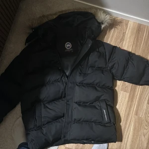 Svart dunjacka från Canada Goose (står storlek Xl men den är M) - Svart dunjacka från Canada Goose med puffy design och huva med pälsdetalj. Jackan har klassisk logga på ärmen, stora fickor med logotyp och dragkedja samt knappar framtill. Perfekt för kalla vinterdagar och riktigt snygg streetstil. (den passar mig och jag är 180 så jag vet inte rintigt om det är barn xl men jag antar det. 60-65kg