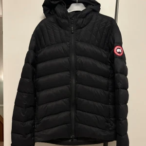  Canada Goose dunjacka - Säljer en svart dunjacka från Canada Goose med huva och klassisk logga på ärmen. Jackan har quiltad design, dragkedja framtill och är perfekt för kalla dagar. Fylld med dun och har en snygg, stilren look som funkar till alla dina outfits. Kvitto finns