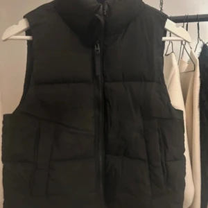 Svart dunväst från Stradivarius - Svart dunväst från GinaTricot i storlek XS. Västen har hög krage, quiltad design och dragkedja framtill. Perfekt att slänga över en hoodie för extra värme. Enkel och stilren look som funkar till många outfits.