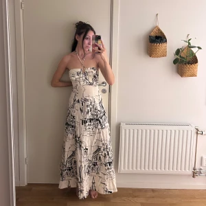 Ny Vit långklänning med svart mönster från Zara - Ny, Säljer en vit långklänning från Zara i storlek XS. Klänningen är axelbandslös med halterneck och har en snygg, figurnära passform upptill och utsvängd kjol. 