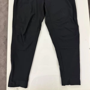 Svarta Nike träningsbyxor  - Svarta Nike träningsbyxor med elastisk midja och dragsko. Byxorna har smal passform, dragkedjeförsedda fickor och diskret Nike-logga på benet. Perfekta för sport eller chill, tillverkade i mjukt syntetmaterial.