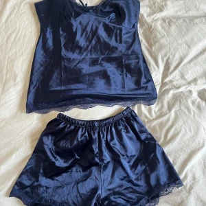 Satinpyjamas  - Snygga mörkblå shorts i satin med elastisk midja och dekorativ spetskant längst ner. Perfekta för chill hemma eller som pyjamas. Materialet är glansigt och mjukt mot huden, och färgen är djup marinblå. Matcha med linne för en komplett look.
