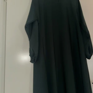 Svart långklänning med vida ärmar - Säljer en grön långklänning från Ludeecollection med rund halsringning och diskret söm framtill. Klänningen har vida, långa ärmar är gjord i ett mjukt, lätt material som faller snyggt. Perfekt för dig som gillar enkel och stilren design.