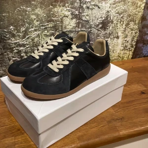 Maison Margiela svarta sneakers - Maison Margiela sneakers i svart skinn och mocka med beige snörning och gummisula. Klassisk och stilren design med rund tå och kontrasterande ljus innersida. Kommer med originalkartong och dustbags. PRIS KAN DISKUTERAS!