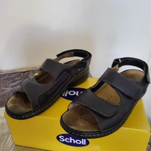 Svarta sandaler från Scholl i skinn - Svarta sandaler från Scholl med bekväm platt sula och öppna tår. Tillverkade i skinn med kontrasterande vita sömmar och justerbara kardborreband. Insidan är mjuk och fotbädden har små perforeringar för extra komfort. Perfekt för varma dagar.