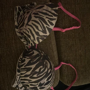 Zebramönstrad Calvin Klein Bh - Säljer en snygg bh från Calvin Klein med zebramönster i svart, grått och beige. Axelbanden är starkt rosa och justerbara för perfekt passform. Kuporna är formade och har en klassisk hakknäppning bak. Perfekt för dig som gillar djurmönster och färg! Det är en b - kupa men vet inte mer då jag klippt av lappen.