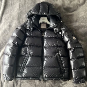 Svart pufferjacka från Moncler - Säljer en svart pufferjacka från Moncler med glansig finish och huva. Jackan har två sidofickor med dragkedja, quiltad design och Moncler-logga på ärmen. Perfekt för kalla dagar och har en modern, oversized look.
