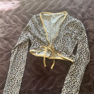 Leopardmönstrad croppad bolerotopp - Säljer en croppad bolerotopp med leopardmönster och guldfärgade kanter. Toppen har långa ärmar och knyt framtill för en snygg och trendig look. Perfekt för dig som vill sticka ut med djurprint och coola detaljer.