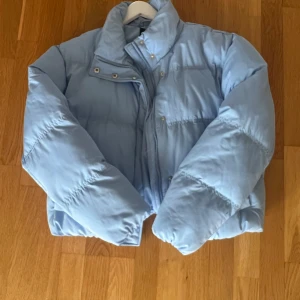 Ljusblå pufferjacka med dragkedja - Säljer en ljusblå pufferjacka med hög krage och dragkedja framtill. Jackan har en quiltad design och tryckknappar vid halsen. Perfekt för kalla dagar och har en loose passform som ger en chill vibe.