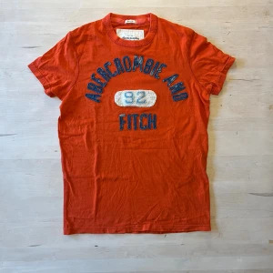 Orange Abercrombie & Fitch t-shirt S - Snygg orange t-shirt från Abercrombie & Fitch med blått lappat tryck och siffran 92 i vitt på bröstet. Klassisk rund hals och korta ärmar. Materialet är mjuk bomull och passformen är slim muscle fit. Perfekt för dig som gillar casual stil. Storlek S, fint skick.