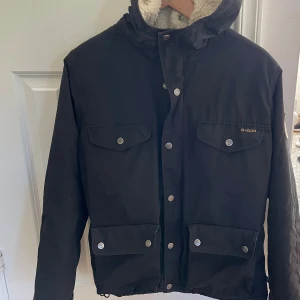 Svart Fjällräven teddyjacka XS - Svart teddyjacka från Fjällräven i storlek XS med G-1000-tyg och mysigt vitt fleecefoder. Jackan har huva, fyra fickor med tryckknappar och snygga detaljer. Perfekt för kalla dagar och har en cool outdoor-vibe.