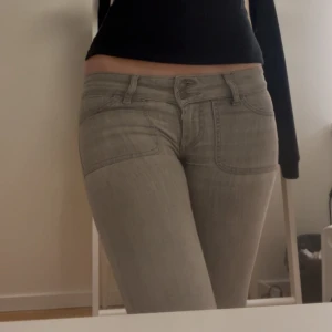 Grå jeansbyxor med låg midja - Säljer ett par gråa lågmidjade jeans botchutt. Byxorna har framfickor och bakfickor med lock och knappdetalj. Materialet är klassisk jeans och passformen är normal, köpte dessa på plick men de var tyvärr försmå