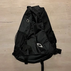 Svart Oakley sling ryggsäck med hexagonmönster - Säljer en svart ryggsäck från Oakley med coolt hexagonmönster och vit broderad logga. Väskan har meshfickor på sidorna, justerbara remmar och en unik asymmetrisk form. Perfekt för dig som vill ha en sportig och funktionell look. Ena remmen har lossnat lite i sömmen vilket syns på sista bilden. Utöver det i väldigt fint skick.