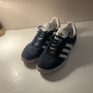 Adidas Gazelle sneakers i marinblå mocka - Säljer ett par klassiska Adidas Gazelle sneakers i marinblå mocka med vita detaljer. Skorna har vit plös och häl, samt marinblå snörning och platt sula. Perfekta för en stilren vardagslook. Priset går att diskutera.