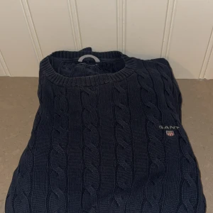 Mörkblå kabelstickad tröja från GANT - Kabelstickad mörkblå tröja från GANT med rund halsringning och broderad logga på bröstet. Tröjan har lång ärm och klassisk passform, materialet är mjukt och skönt stickat. Inget fel med tröjan