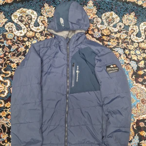 Blå pufferjacka från Sail Racing - Blå pufferjacka från Sail Racing med huva och snygga detaljer på bröstet och ärmen. Jackan har dragkedja framtill, en bröstficka med zip och fodrad insida i grått. Perfekt för kalla dagar och har en sportig look med logga på huvan och bröstet.