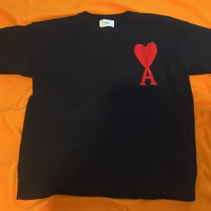 Svart stickad tröja från Ami Paris - Svart stickad tröja från Ami Paris med röd hjärtlogga och bokstaven A på bröstet. Klassisk rund halsringning och ribbade muddar. Perfekt för dig som vill ha en stilren men ändå unik look.