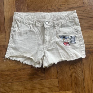 Vita shorts med brodyr från H&M - Säljer ett par vita shorts från H&M med råa kanter och coola broderade detaljer som ett bi, hjärta och texten 'Whatever Forever' på framsidan. Klassisk femficksmodell i bomull med lite stretch för skön passform. Perfekta för sommaren!