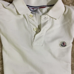 Beige Moncler piké med logga - Beige  pikétröja från Moncler i storlek S. Klassisk krage, två knappar framtill och Moncler-logga broderad på bröstet. Materialet är mjuk bomull och kragen har en snygg detalj med röd och marinblå rand på insidan. Perfekt för dig som gillar stilrena märkesplagg.