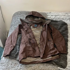 Vindjacka Helly Hansen - Brun vindjacka från Helly Hansen i storlek XS (stor). Jackan har huva, Helly Tech-teknologi,  meshfoder på insidan och är hyfsat lång. Perfekt för blåsiga dagar!