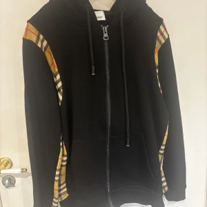 Burberry tröja  - Svart hoodie från Burberry med klassiska rutiga paneler i beige, rött och svart längs axlar och sidor. Tröjan har dragkedja framtill, huva med snörning och två fickor. Perfekt för dig som vill ha en stilren men ändå ikonisk look. Storlek xs men passar S