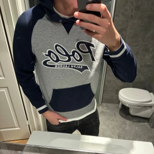 Grå och marinblå hoodie från Polo Ralph Lauren - Säljer en snygg hoodie från Polo Ralph Lauren i grått och marinblått med stort Polo-tryck på bröstet. Riktigt bra skick och inga defekter. Hoodien är i storlek S och är för liten på mig som är 188cm. Hör av er vid funderingar:)