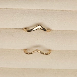 Två guldiga V-formade ringar - Säljer två stilrena guldiga ringar med V-form. Den ena är slät och enkel medan den andra är dekorerad med små gnistrande stenar längs hela ovansidan. Perfekta att bära tillsammans eller var för sig för en trendig look.